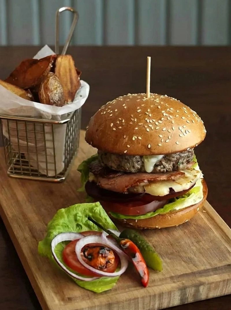 Hamburger i lartë me djathë në një tavolinë të ngrohtë, me patate anash